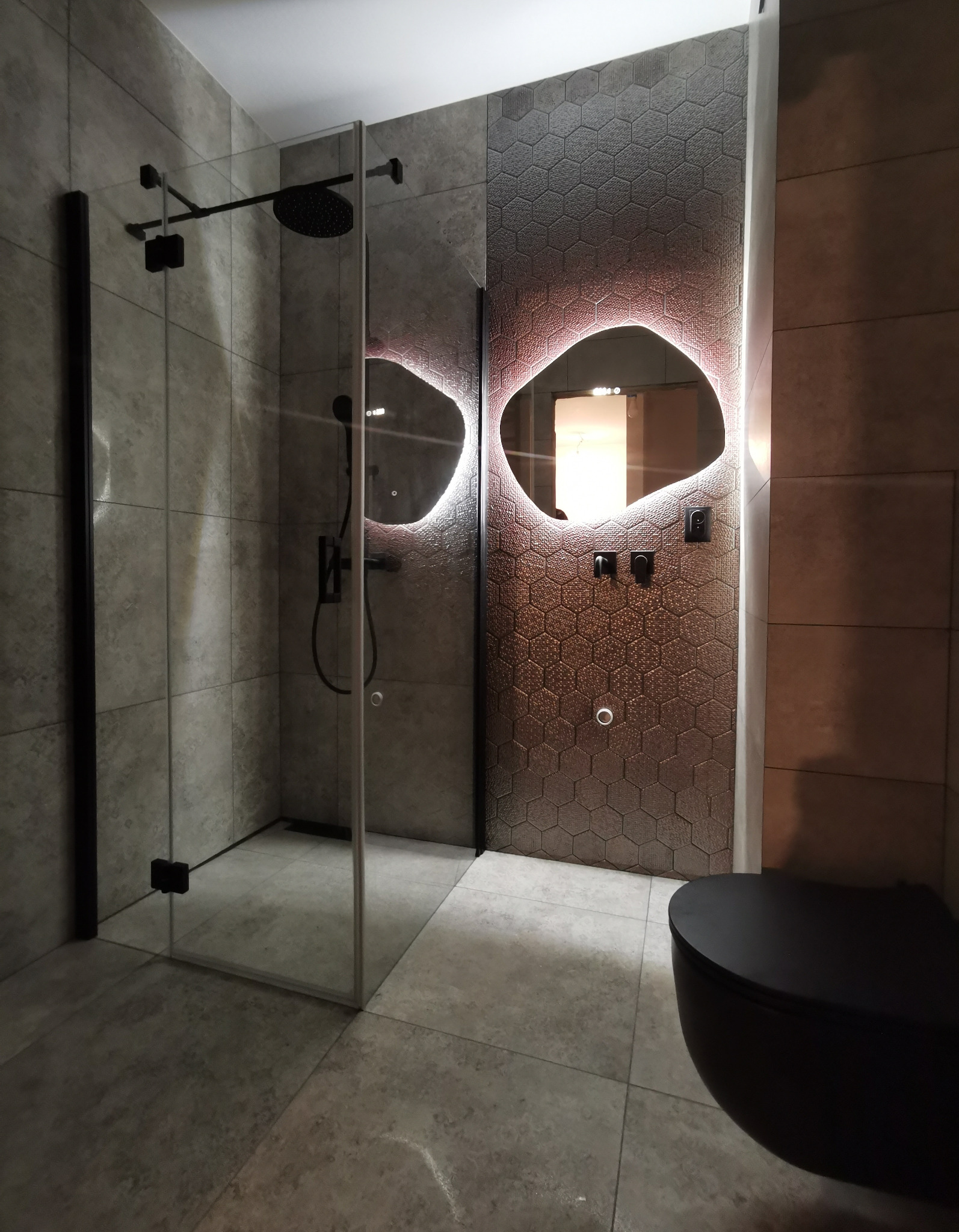 Nowoczesna łazienka z kabiną prysznicową typu walk-in z czarnymi elementami, heksagonalnymi płytkami z podświetleniem LED wokół lustra i wiszącą czarną toaletą.