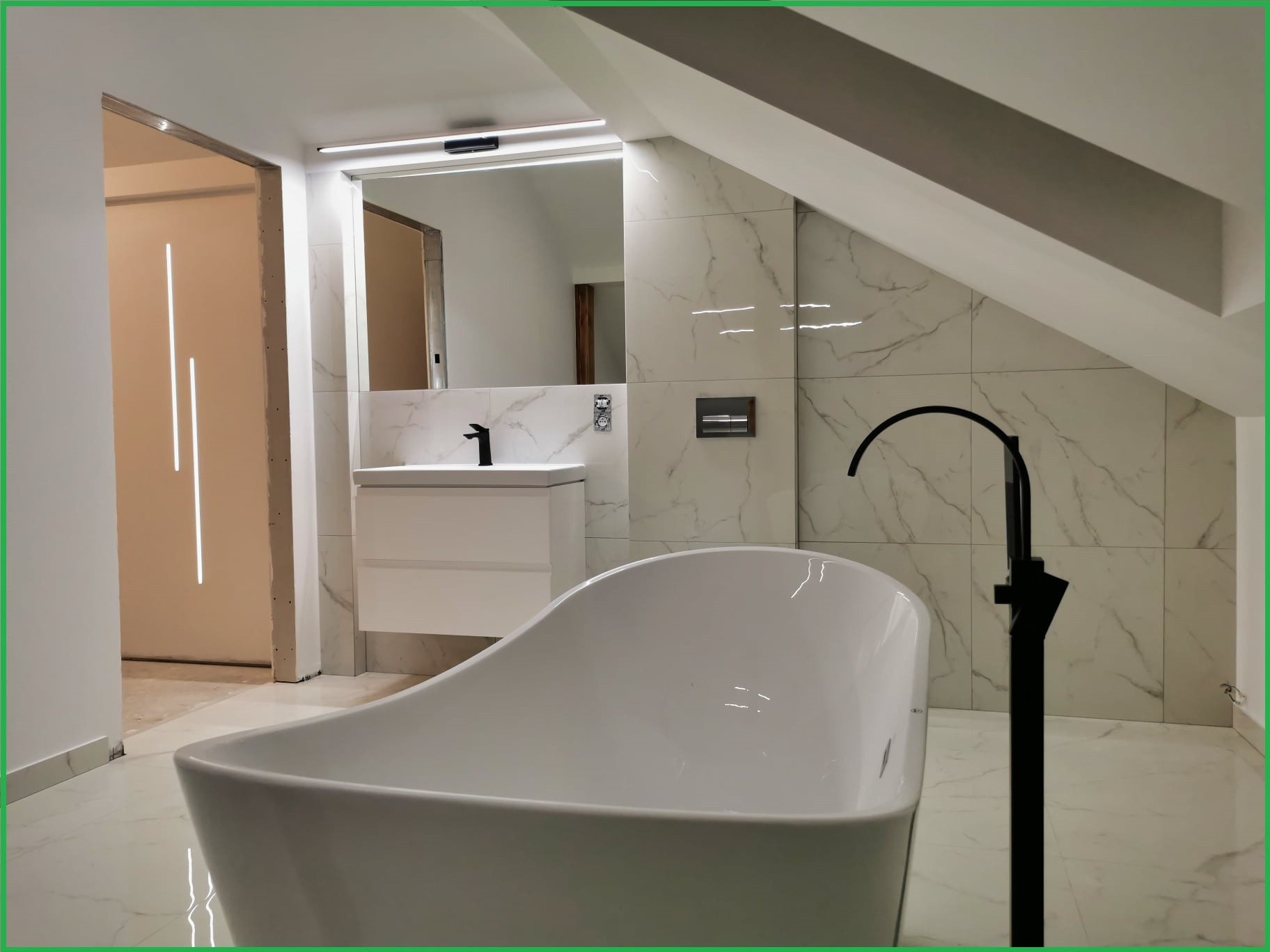 Nowoczesna łazienka na poddaszu z wolnostojącą białą wanną, czarną armaturą i marmurowymi płytkami, widok na umywalkę z podświetlanym lustrem i otwarte drzwi.