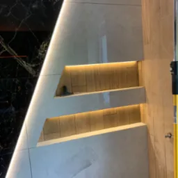 Nowoczesna łazienka z czarnym marmurem i jasnymi płytkami, oświetlone półki w ścianie, widoczny fragment narzędzia.