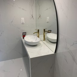 Usługi remontowe - Biała umywalka nablatowa z baterią w kolorze złotym na blacie z imitacji marmuru, owalne lustro z czarną ramą, ściany i podłoga wyłożone płytkami imitującymi marmur, widoczne niedokończone...