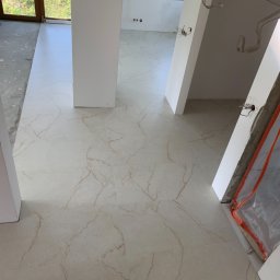 Usługi remontowe - Jasne, marmurkowe płytki podłogowe z brązowymi żyłami, świeżo ułożone w surowym wnętrzu domu, z widocznymi instalacjami elektrycznymi i oknem w tle.