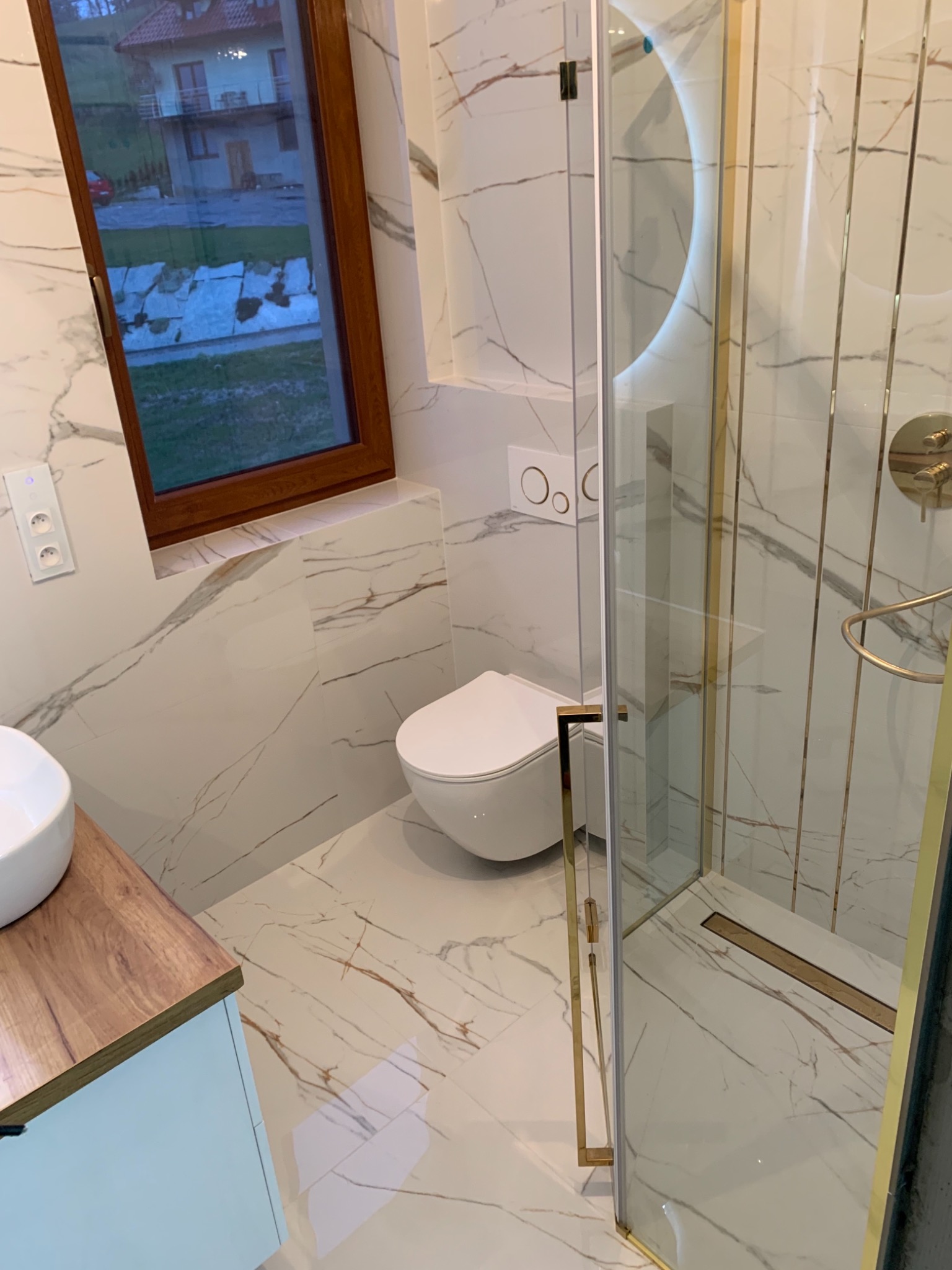 Nowoczesna łazienka z marmurowymi płytkami, kabiną prysznicową ze złotymi elementami, podwieszaną miską WC, drewnianym blatem umywalki i widokiem z okna na dom.