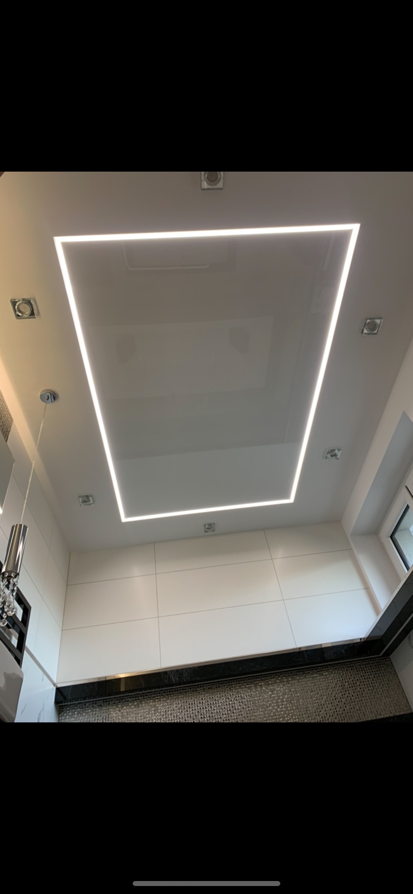 Biały sufit podwieszany z podświetleniem LED w kształcie prostokąta, widoczne punktowe oświetlenie i fragment ściany wyłożonej białymi płytkami.