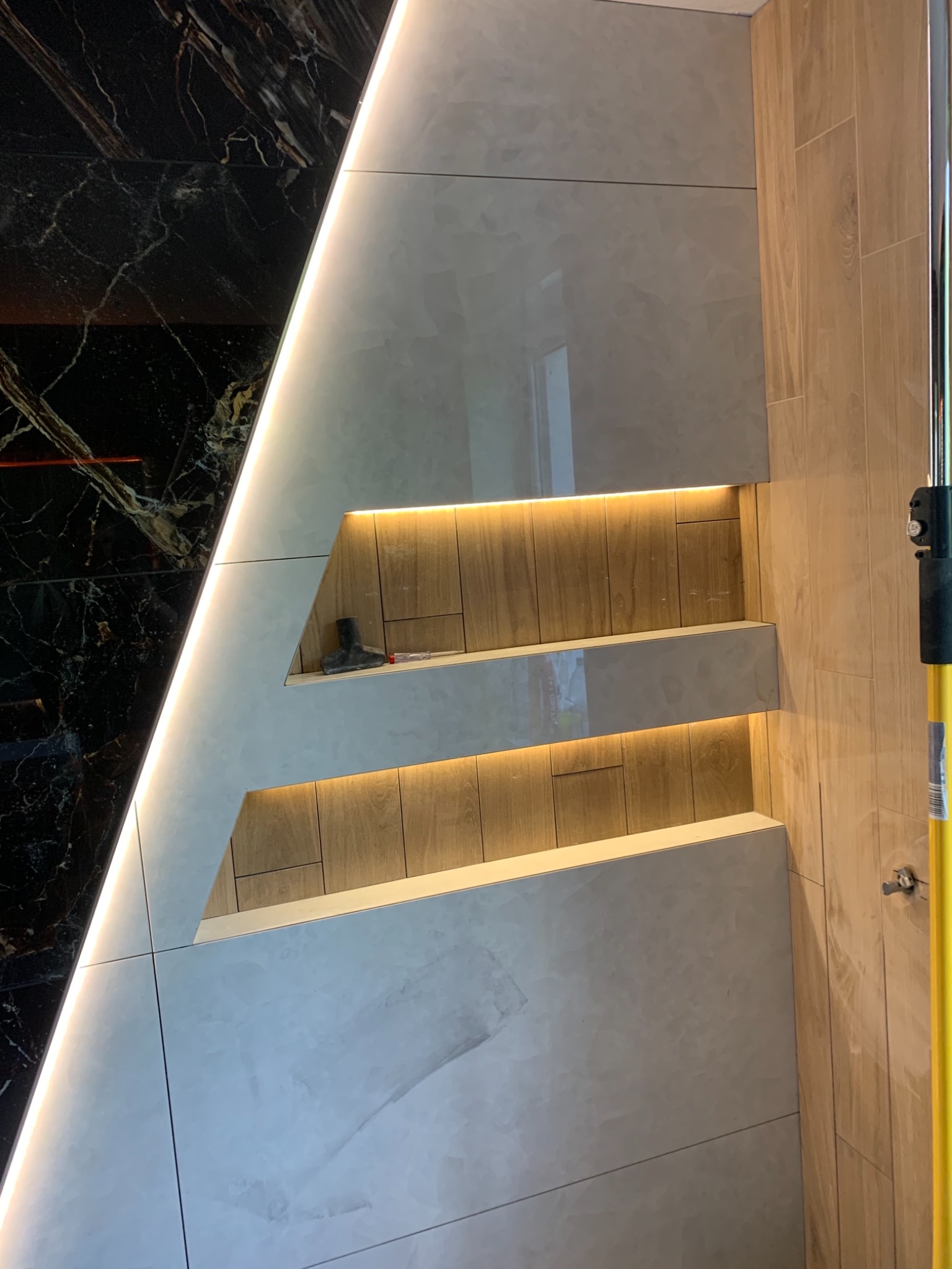 Nowoczesna łazienka z czarnym marmurem i jasnymi płytkami, oświetlone półki w ścianie, widoczny fragment narzędzia.