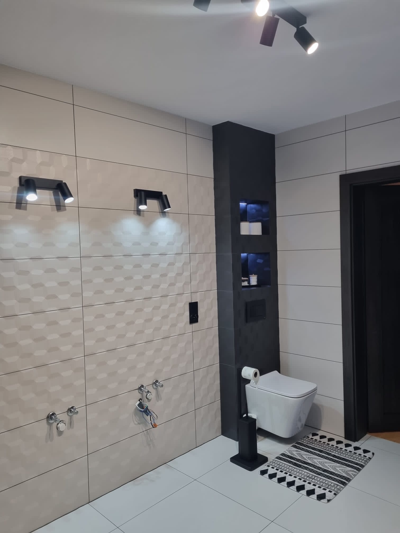 Nowoczesna łazienka z beżowymi płytkami o trójwymiarowej strukturze, podwieszana toaleta z czarnym akcentem w postaci wnęki z półkami i czarnym uchwytem na papier toaletowy, oraz czarne oświetlenie...