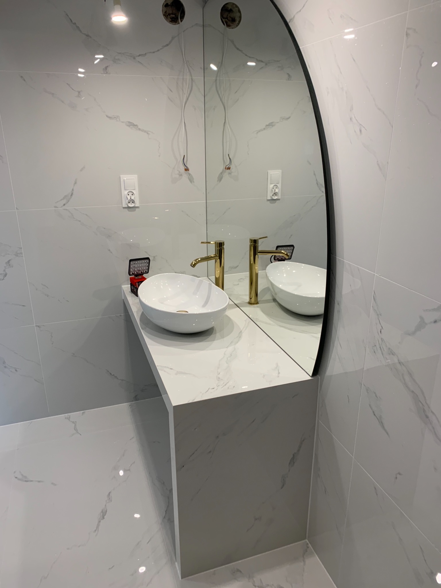 Biała umywalka nablatowa z baterią w kolorze złotym na blacie z imitacji marmuru, owalne lustro z czarną ramą, ściany i podłoga wyłożone płytkami imitującymi marmur, widoczne niedokończone...