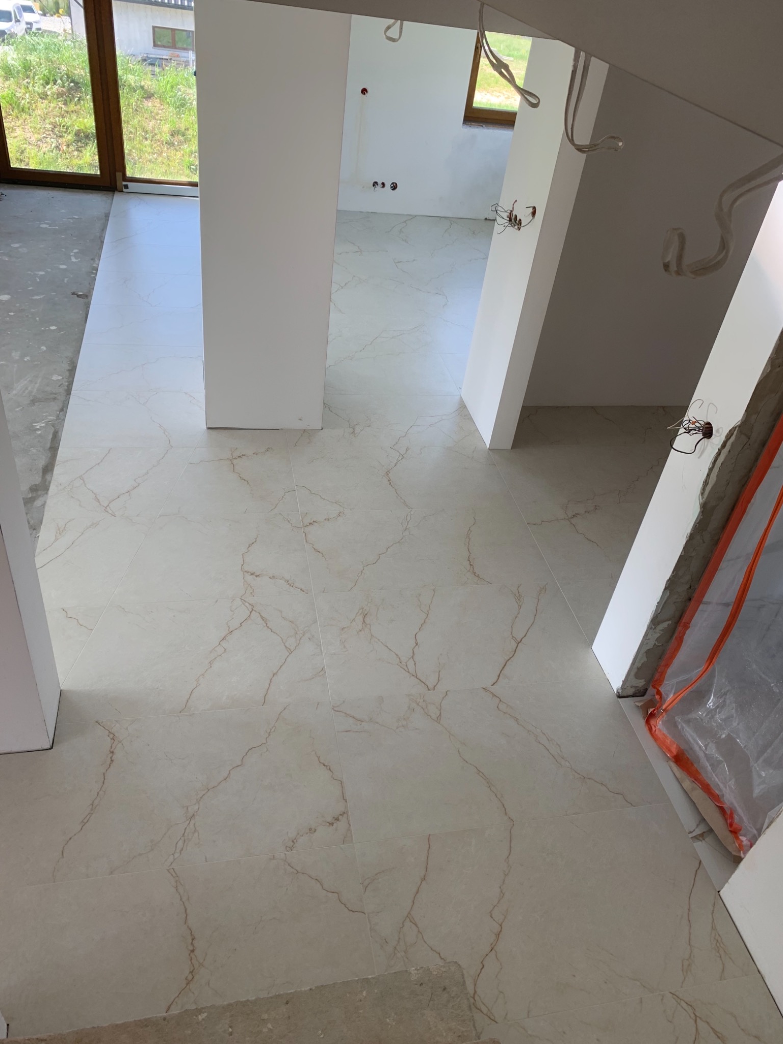 Jasne, marmurkowe płytki podłogowe z brązowymi żyłami, świeżo ułożone w surowym wnętrzu domu, z widocznymi instalacjami elektrycznymi i oknem w tle.