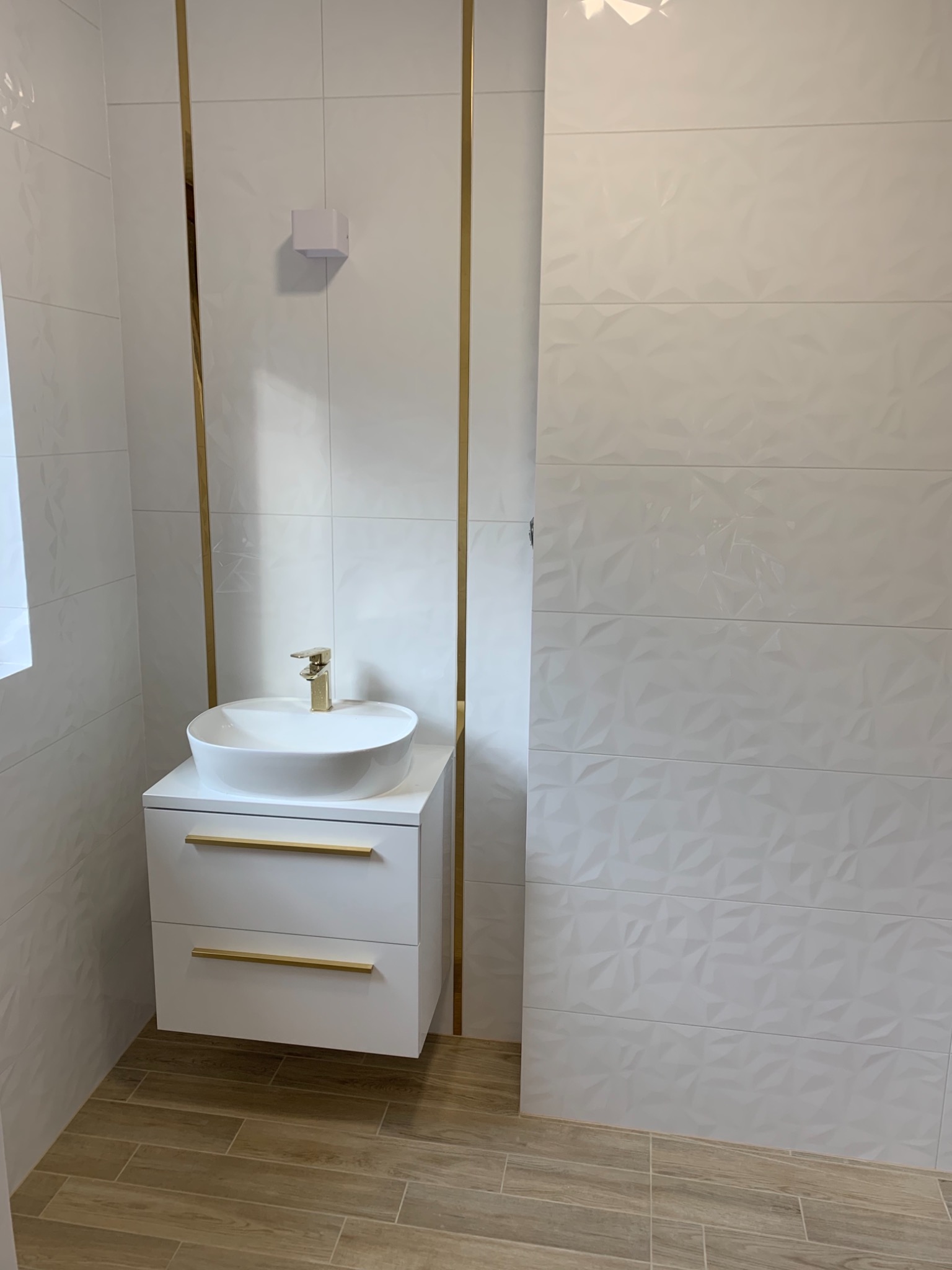 Nowoczesna łazienka z umywalką nablatową, złotą armaturą i dekoracyjnymi białymi płytkami ściennymi z geometrycznym wzorem, drewnopodobna podłoga.