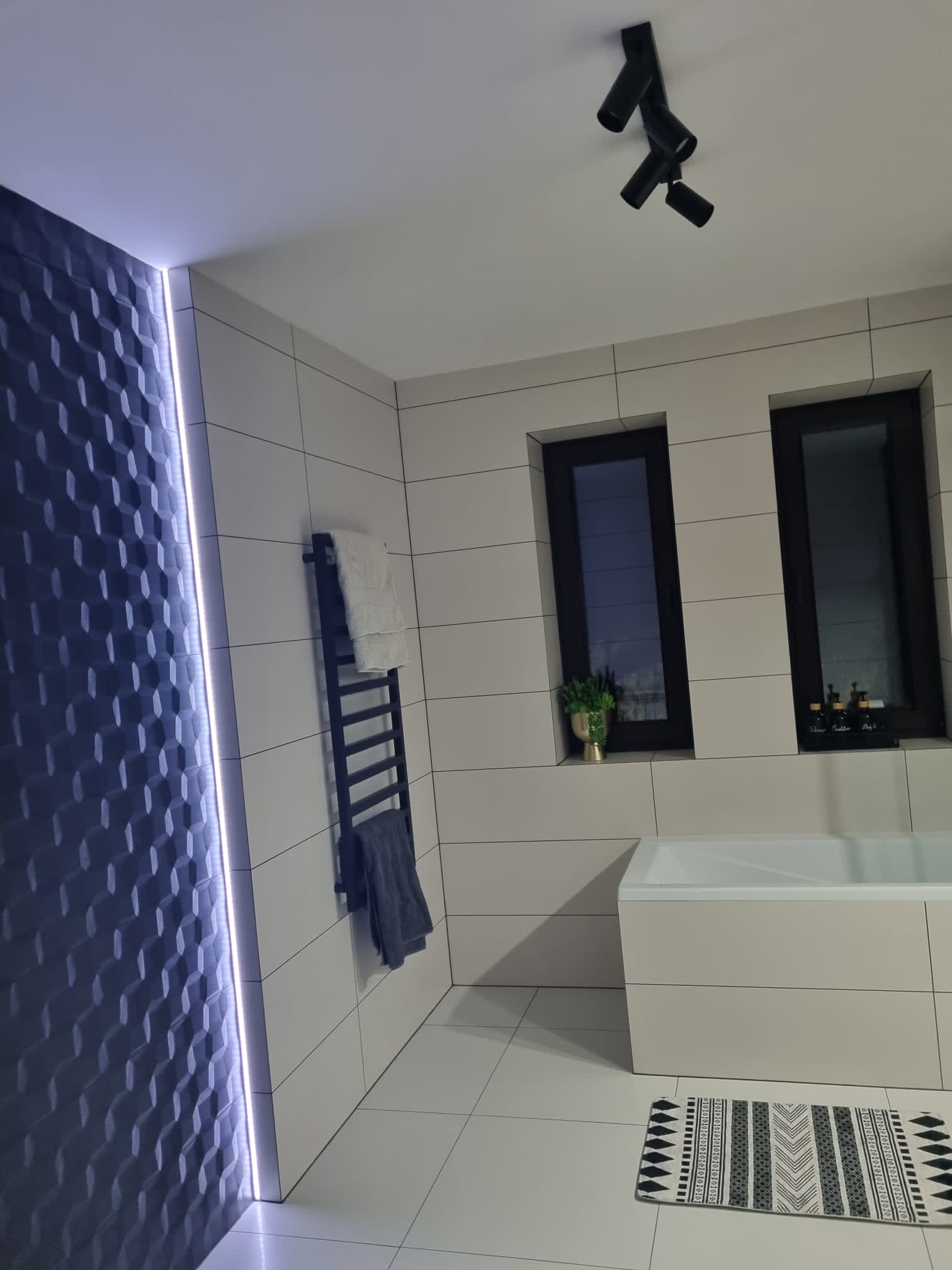Nowoczesna łazienka z geometryczną ścianą podświetloną LED, grzejnikiem drabinkowym na ręczniki, białą wanną i czarnymi ramami okiennymi z dekoracjami.