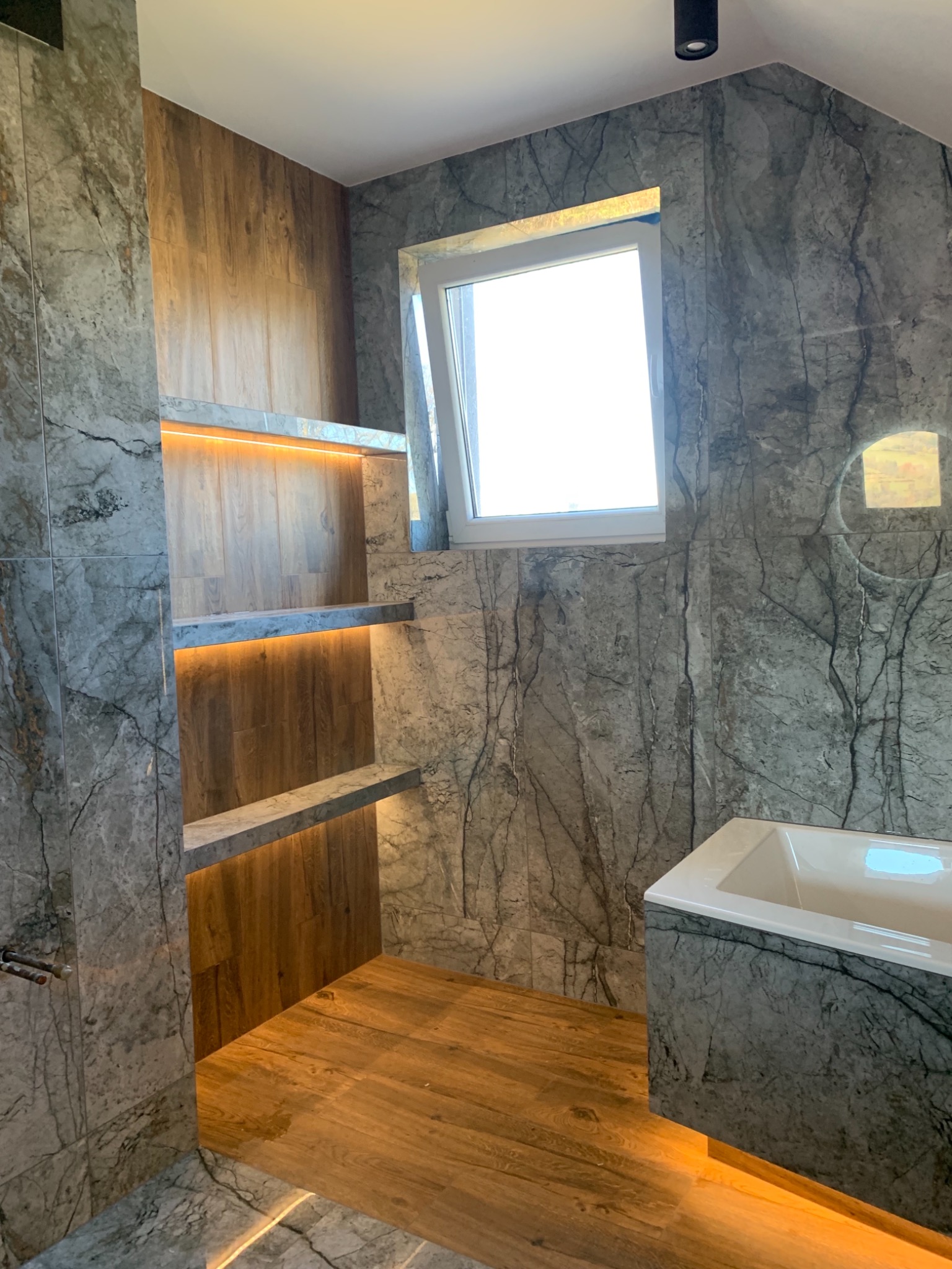 Nowoczesna łazienka z szarą marmurową okładziną, drewnianą podłogą, podświetlanymi półkami i wanną, oświetlona punktowym światłem sufitowym.