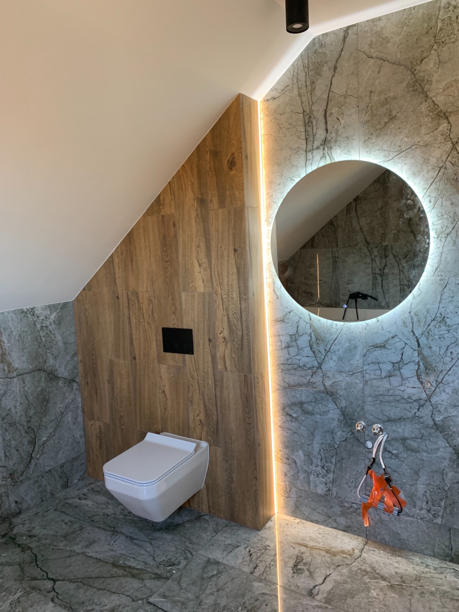 Nowoczesna łazienka na poddaszu z marmurowymi płytkami, podświetlanym lustrem i wiszącą toaletą, wykończona drewnianą boazerią.