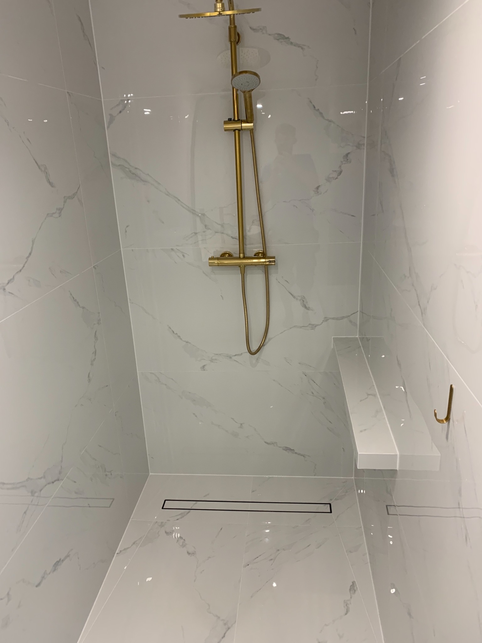 Nowoczesny prysznic typu walk-in z białymi płytkami imitującymi marmur, złotą armaturą i odpływem liniowym.