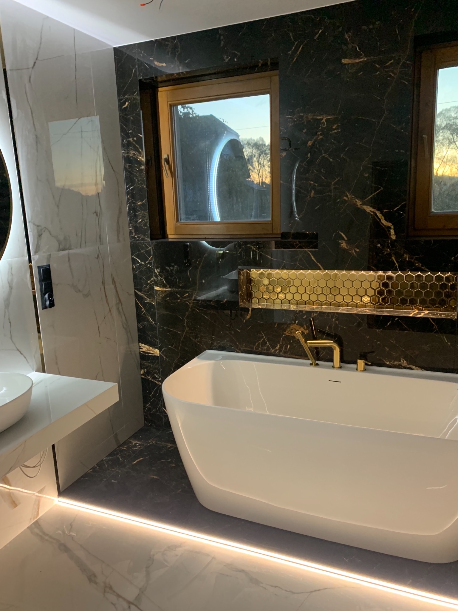 Nowoczesna łazienka z wolnostojącą białą wanną, czarnymi marmurowymi płytkami ze złotymi żyłami, złotą mozaiką heksagonalną nad wanną i podświetleniem LED przy podłodze.