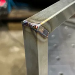 Zbliżenie na spawany narożnik ramy z profili nierdzewnych, widoczne ślady spawania i opalizujące kolory na spoinie.