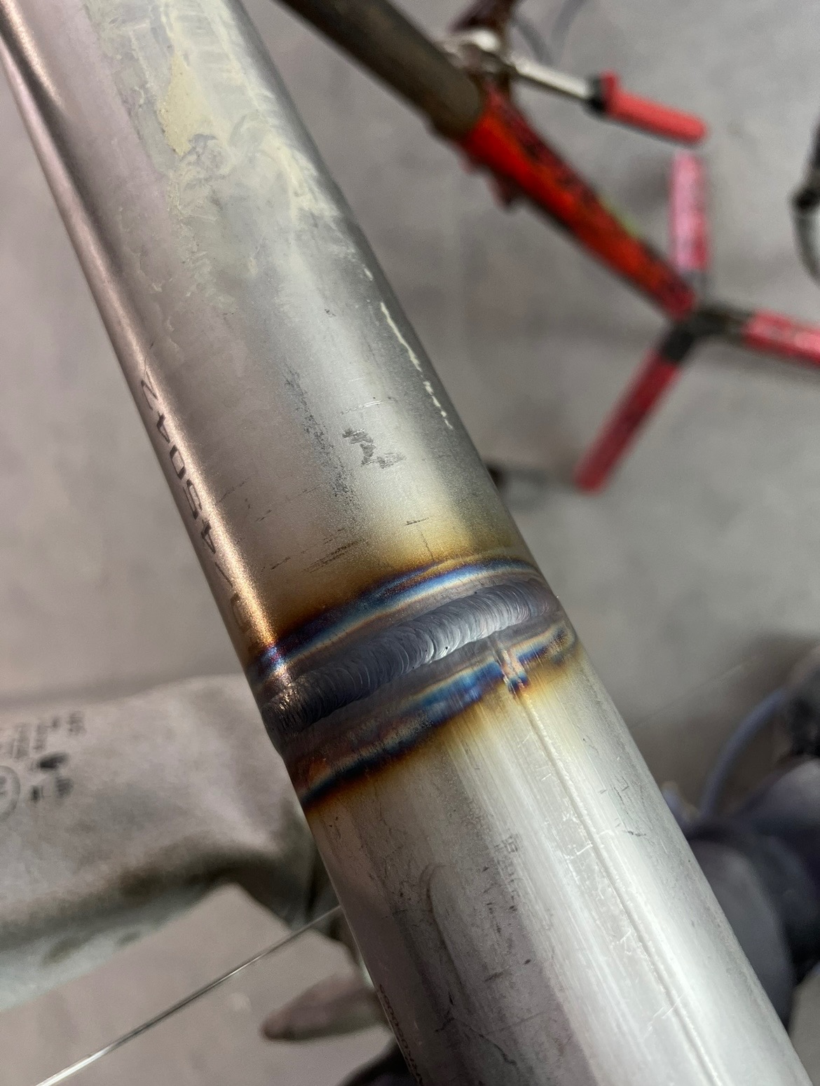 Szczegółowe zbliżenie na spawany szew na rurze ze stali nierdzewnej, widoczne charakterystyczne zabarwienia termiczne i precyzyjne ułożenie spoiny. W tle fragment narzędzi warsztatowych.