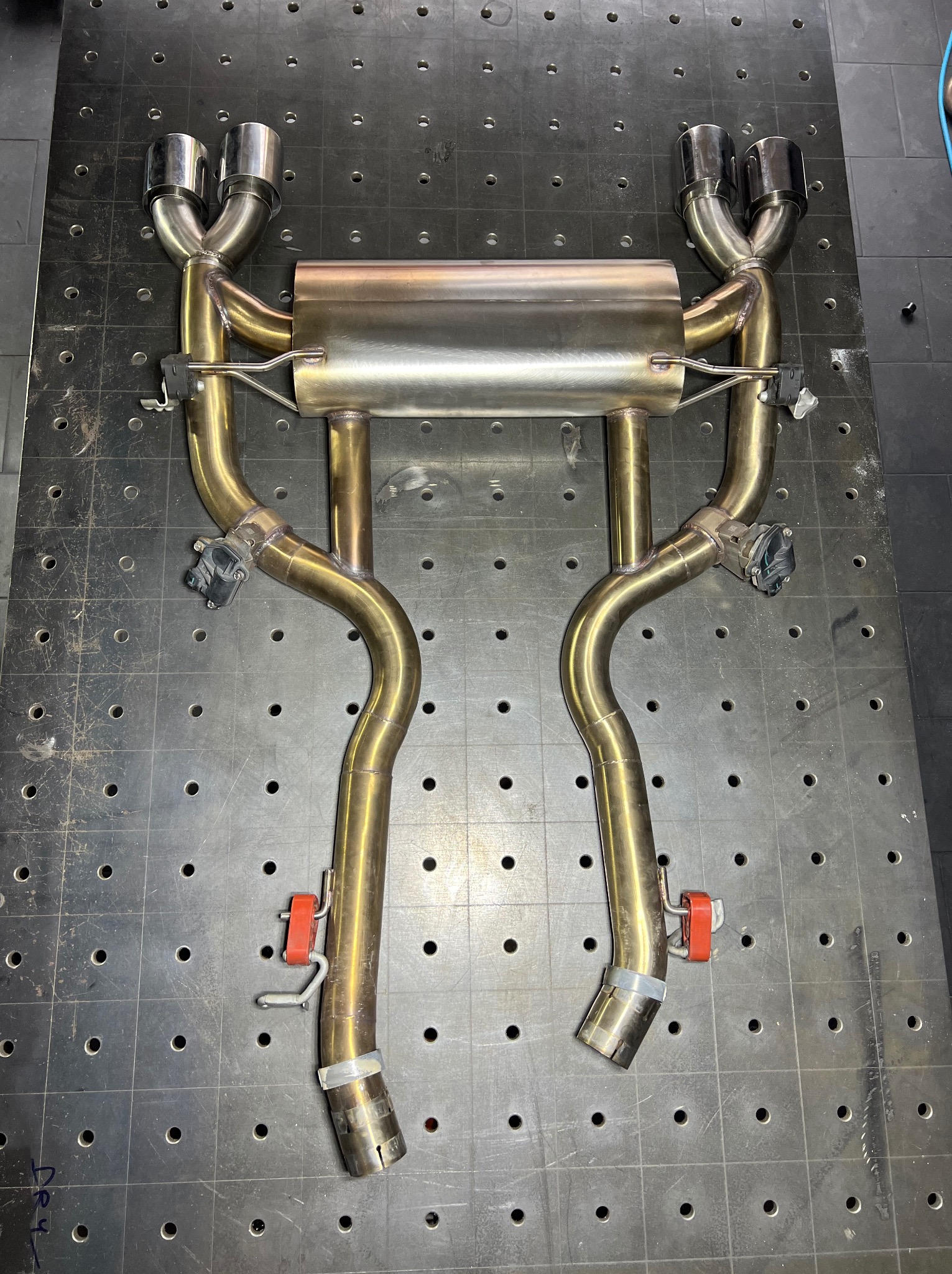 Zespawany układ wydechowy z czterema końcówkami, widoczne spawy i czujniki, leżący na perforowanym stole warsztatowym.
