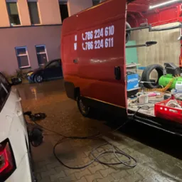 Mobilny serwis oponiarski w akcji: Czerwony van z otwartymi drzwiami, wewnątrz narzędzia i opony, na zewnątrz biały samochód klienta i mokra kostka brukowa.