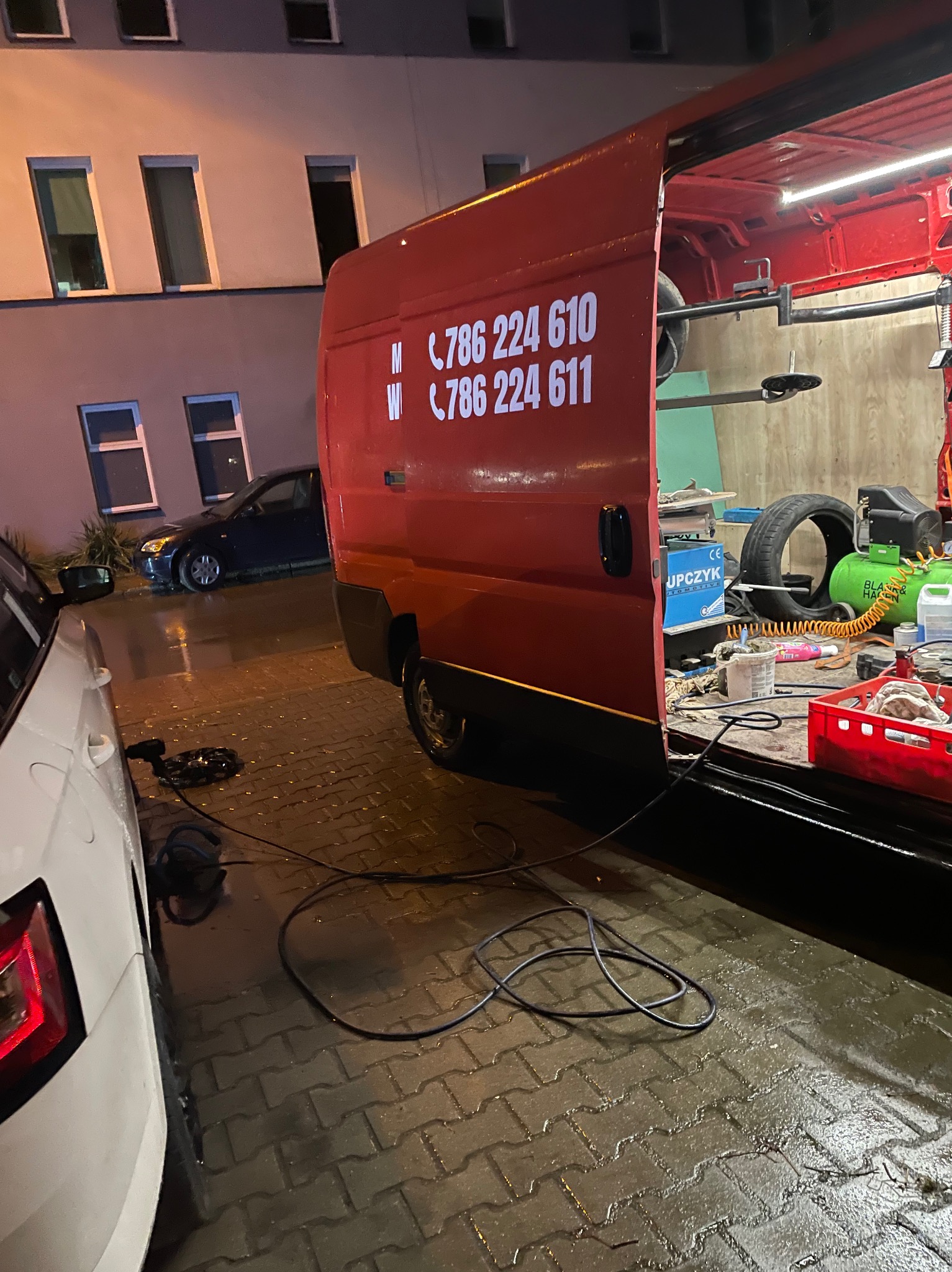 Mobilny serwis oponiarski w akcji: Czerwony van z otwartymi drzwiami, wewnątrz narzędzia i opony, na zewnątrz biały samochód klienta i mokra kostka brukowa.