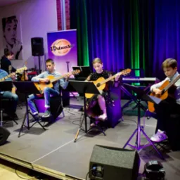Zespół młodych muzyków grających na gitarach klasycznych i basowej na scenie, w tle portret Audrey Hepburn i baner szkoły muzycznej.
