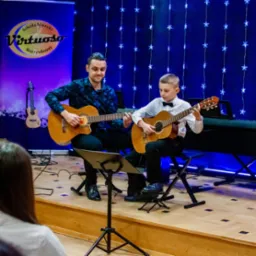 Lekcja gry na gitarze akustycznej: instruktor w kwiecistej koszuli i uczeń w muszce grają na gitarach na scenie z dekoracją świetlną, w tle logo szkoły muzycznej.