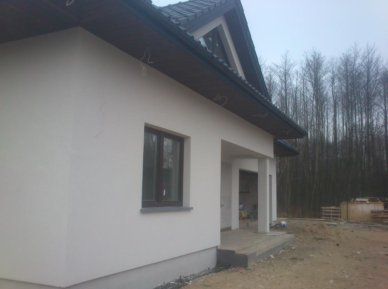 Ocieplony dom jednorodzinny z jasną elewacją i ciemną stolarką okienną, widok z boku na werandę z kolumnami, w tle las bezlistny i palety budowlane.