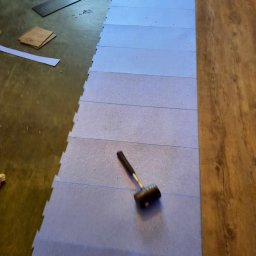 Selfmade Studio Łukasz Jurkowski - Podkład z pianki pod panele podłogowe, gumowy młotek, fragment ułożonych paneli podłogowych, elementy paneli i listew przypodłogowych na podłodze.