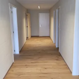 Długi korytarz z nową, jasną podłogą z paneli drewnopodobnych, ściany wykończone w cegiełkę, oświetlenie sufitowe liniowe LED, kilka białych drzwi po obu stronach.