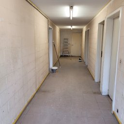 Selfmade Studio Łukasz Jurkowski - Korytarz w trakcie malowania: ściany z bloków pomalowane na jasny kolor, zabezpieczone taśmą malarską przy podłodze i suficie, widoczna drabina, puszka z farbą i wałek malarski.