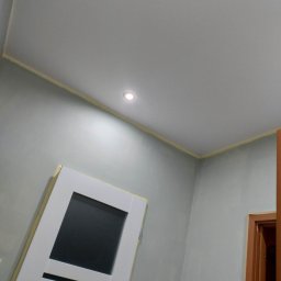 Selfmade Studio Łukasz Jurkowski - Świeżo pomalowane szare ściany z widocznym odcięciem taśmą malarską przy suficie, w tle białe drzwi z czarnym szkłem i fragment ościeżnicy drzwi wewnętrznych.