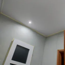 Świeżo pomalowane szare ściany z widocznym odcięciem taśmą malarską przy suficie, w tle białe drzwi z czarnym szkłem i fragment ościeżnicy drzwi wewnętrznych.