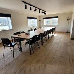 Selfmade Studio Łukasz Jurkowski - Jasna sala konferencyjna z długim stołem, czarnymi krzesłami i drewnianą podłogą. Widoczne okna z roletami i widok na zewnątrz, oraz oświetlenie punktowe na suficie.