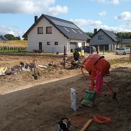 Selfmade Studio Łukasz Jurkowski - Prace ziemne przy budowie domu jednorodzinnego: robotnik z taczką, pomarańczowa betoniarka, worki z zaprawą, widoczne narzędzia i kable na placu budowy, w tle dom z panelami słonecznymi i pochmurne...