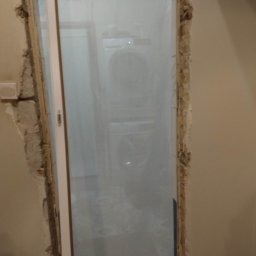 Selfmade Studio Łukasz Jurkowski - Montaż ościeżnicy drzwiowej w trakcie remontu, widoczne uszkodzenia ściany i zabezpieczona folią przestrzeń za drzwiami, fragment nowej podłogi z wzorzystych płytek.