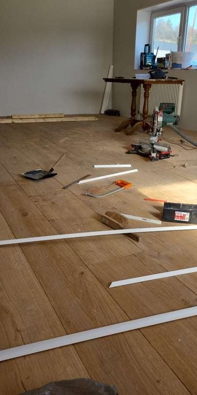 Przygotowanie do montażu listew przypodłogowych na drewnianej podłodze, widoczne narzędzia: piła ukosowa, miarka, pędzel, skrzynka narzędziowa, oraz przycięte listwy.