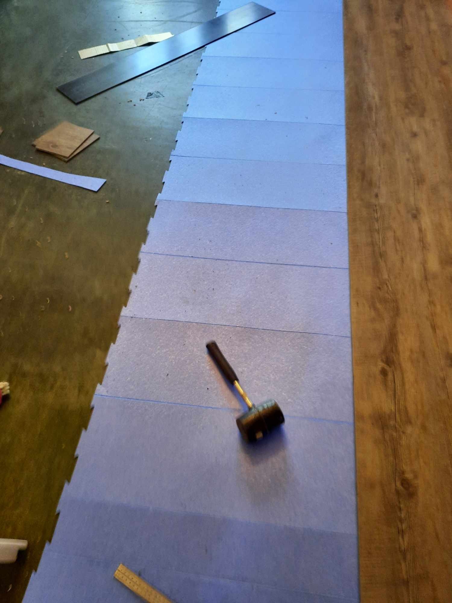 Podkład z pianki pod panele podłogowe, gumowy młotek, fragment ułożonych paneli podłogowych, elementy paneli i listew przypodłogowych na podłodze.