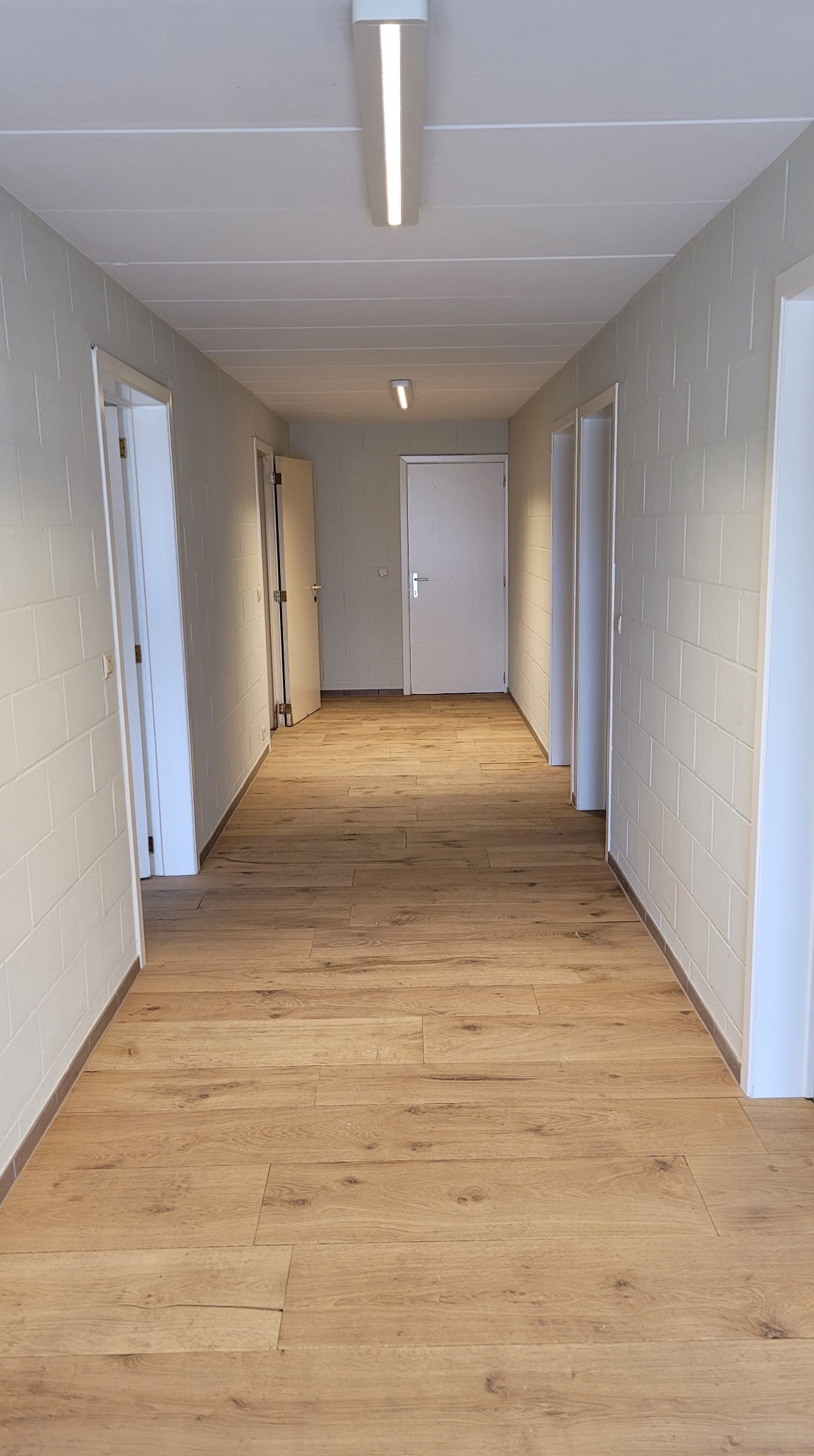 Długi korytarz z nową, jasną podłogą z paneli drewnopodobnych, ściany wykończone w cegiełkę, oświetlenie sufitowe liniowe LED, kilka białych drzwi po obu stronach.