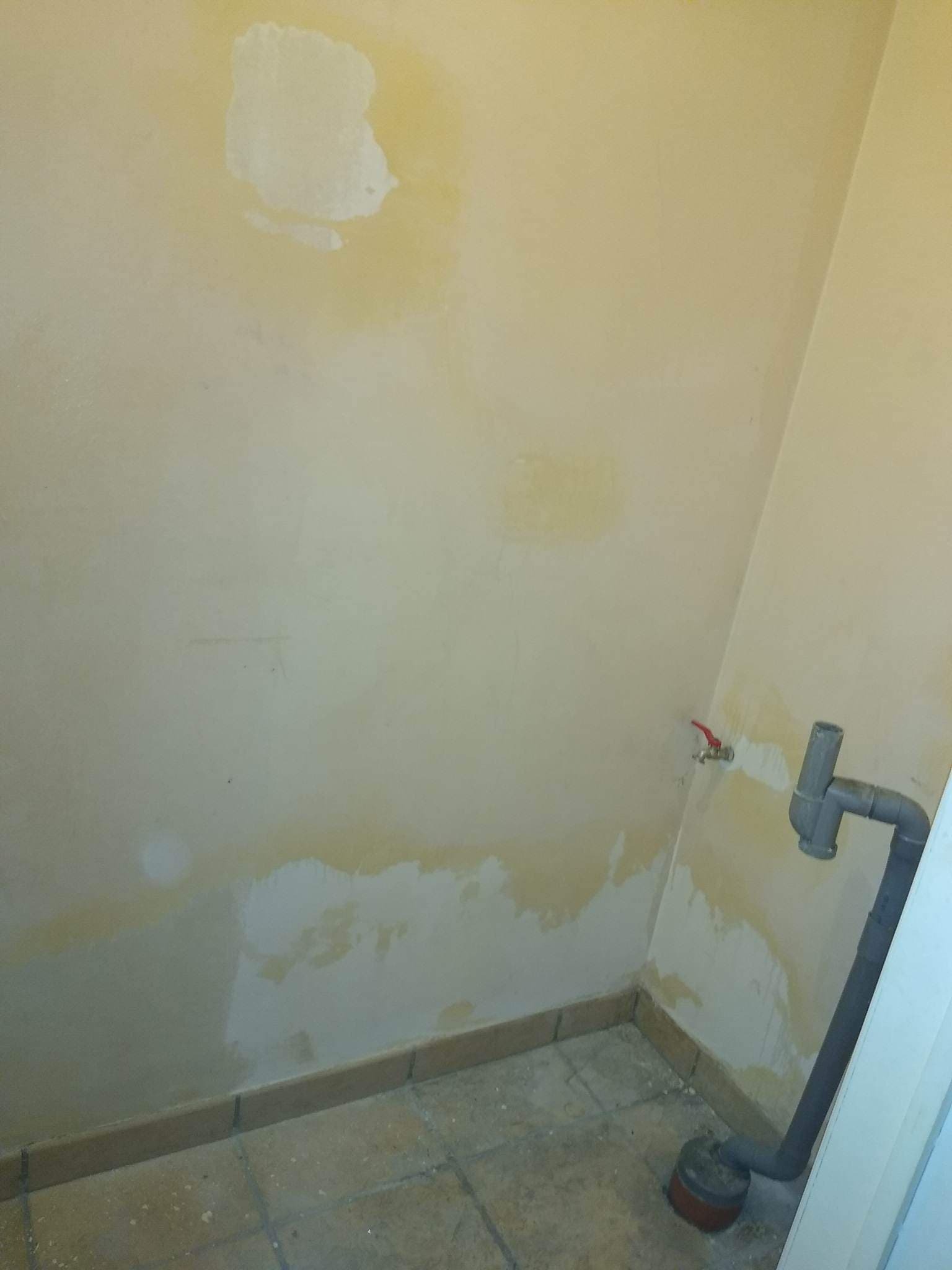 Fragment ściany w trakcie przygotowania do malowania z widocznymi plamami i śladami szpachlowania, szara rura kanalizacyjna przy podłodze i zawór wodny na ścianie.