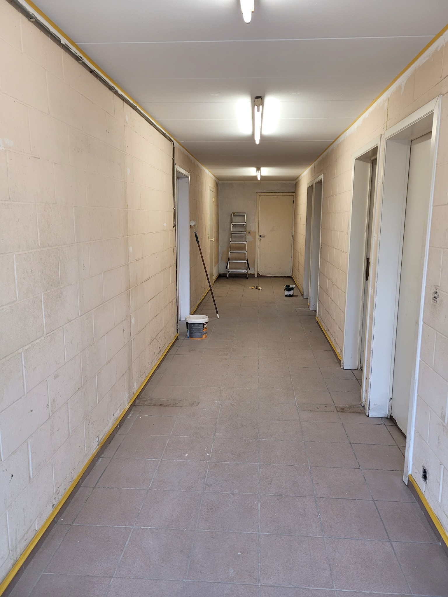 Korytarz w trakcie malowania: ściany z bloków pomalowane na jasny kolor, zabezpieczone taśmą malarską przy podłodze i suficie, widoczna drabina, puszka z farbą i wałek malarski.