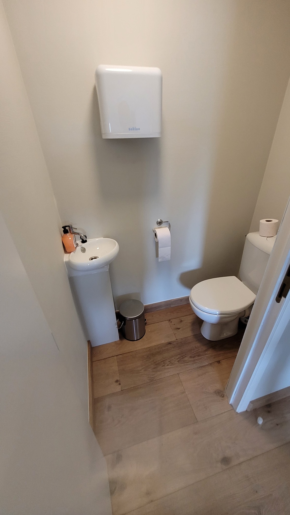 Mała łazienka z drewnianą podłogą, umywalką na postumencie z dozownikiem mydła, toaletą i podajnikiem na ręczniki papierowe marki Sabino.
