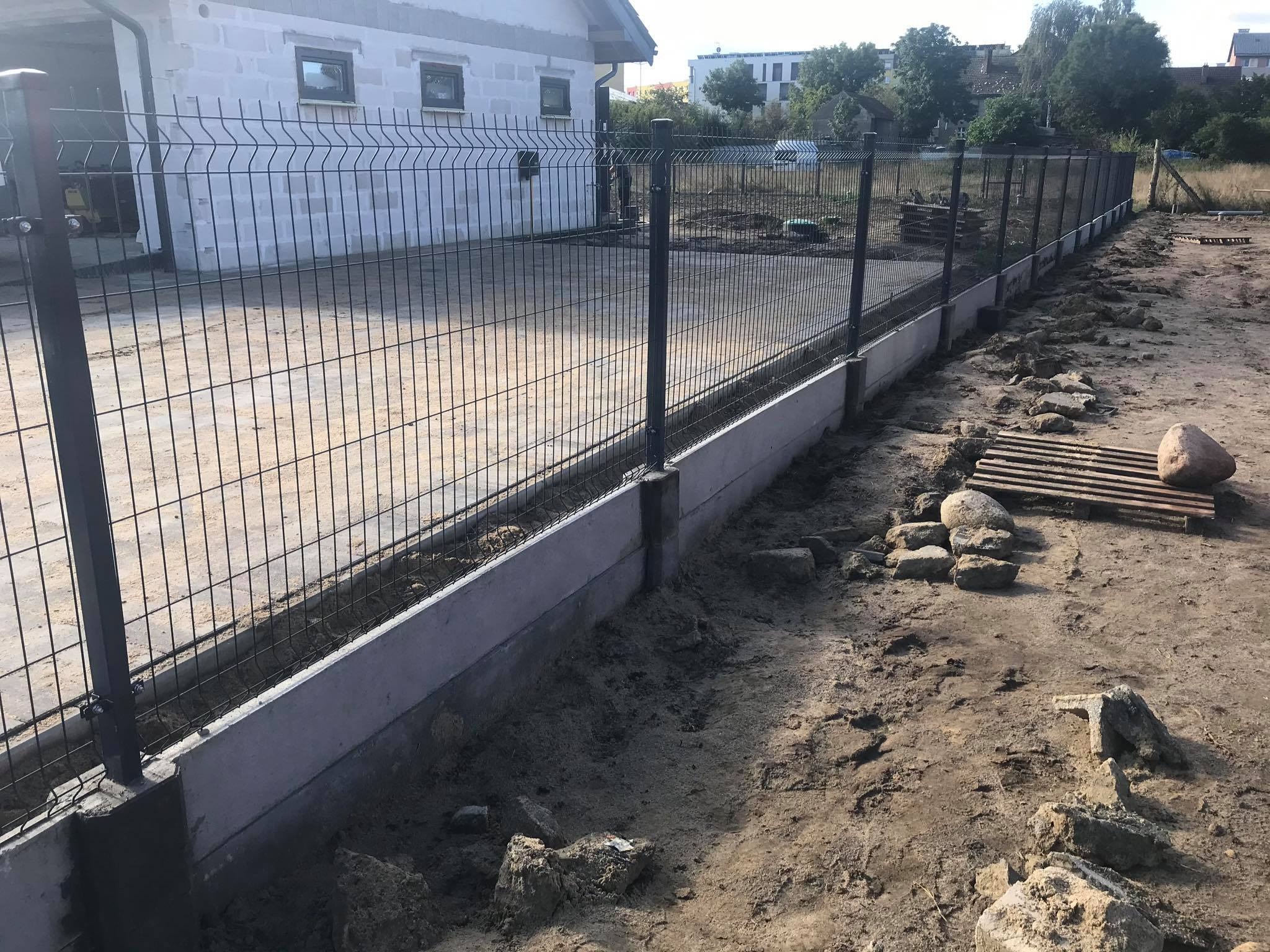 Nowo postawione, stalowe ogrodzenie panelowe z podmurówką betonową, widoczne ślady prac ziemnych, w tle budynek jednorodzinny z oknami.