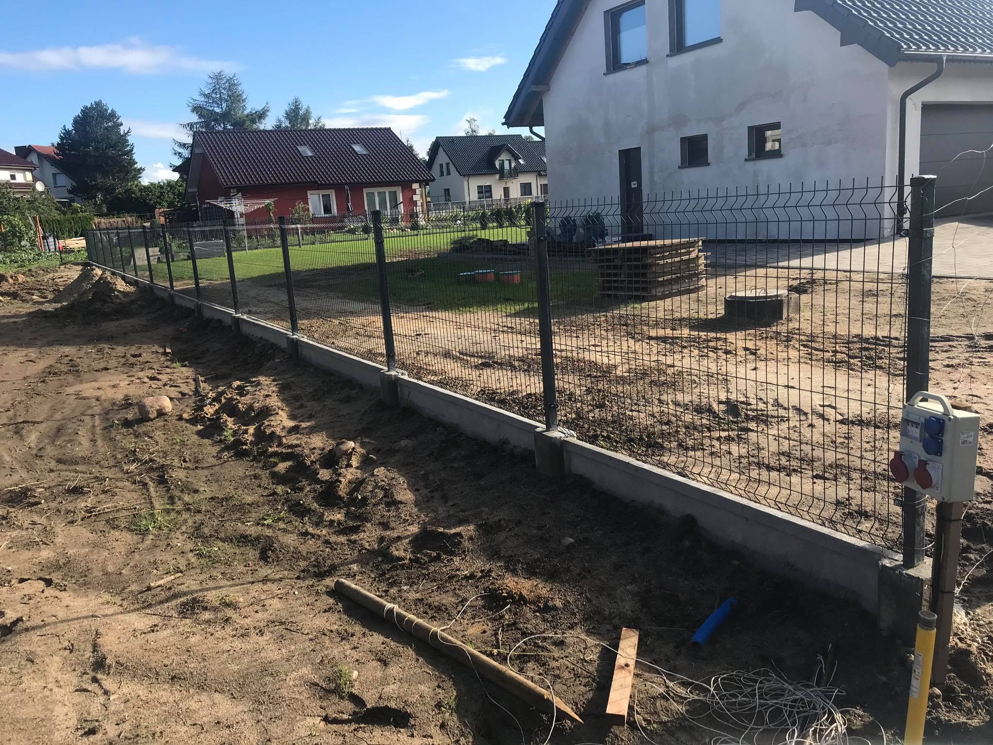 Montaż ogrodzenia panelowego z podmurówką betonową, widoczny fragment ogrodzenia, słupki i siatka, teren wokół w trakcie prac ziemnych, w tle domy jednorodzinne.