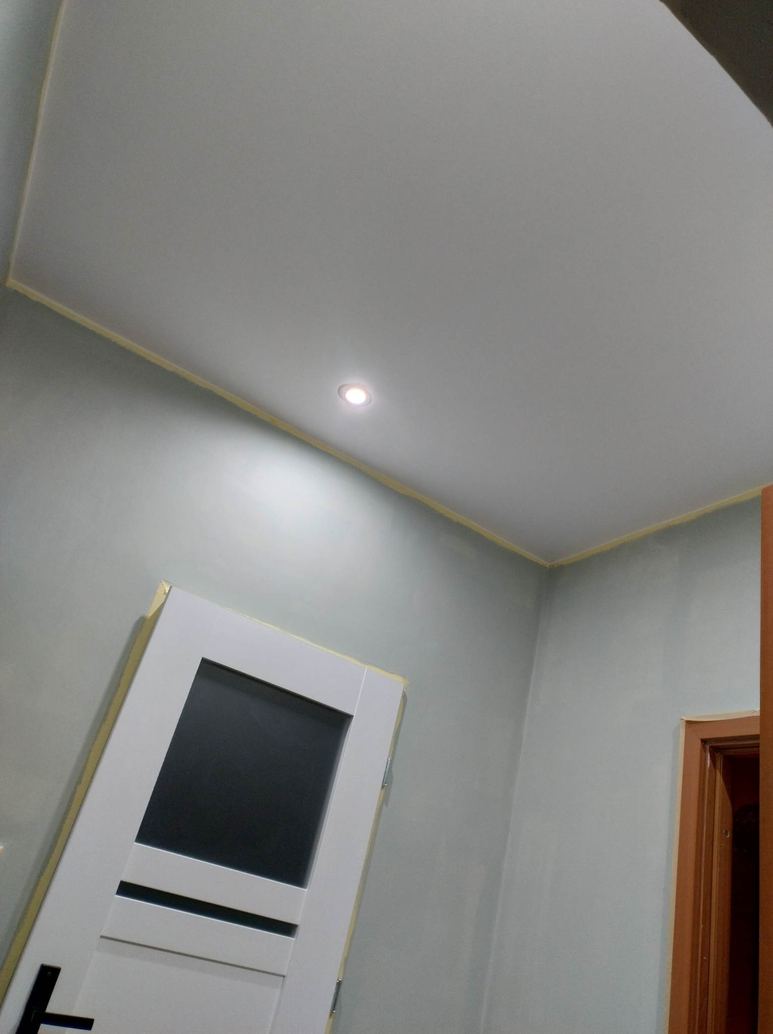 Świeżo pomalowane szare ściany z widocznym odcięciem taśmą malarską przy suficie, w tle białe drzwi z czarnym szkłem i fragment ościeżnicy drzwi wewnętrznych.