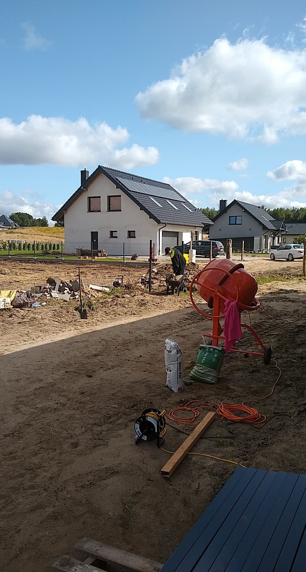 Prace ziemne przy budowie domu jednorodzinnego: robotnik z taczką, pomarańczowa betoniarka, worki z zaprawą, widoczne narzędzia i kable na placu budowy, w tle dom z panelami słonecznymi i pochmurne...