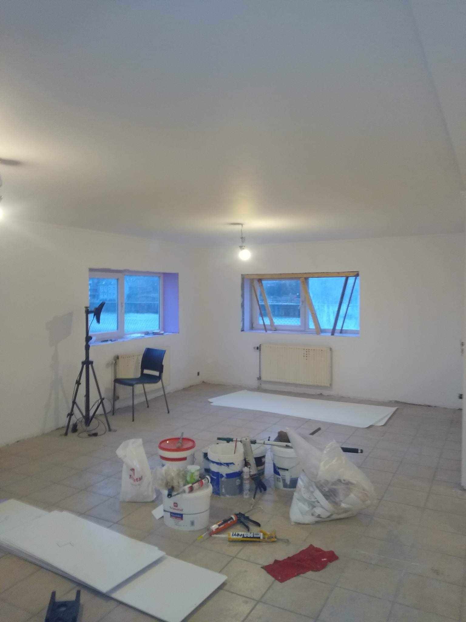 Przygotowania do prac wykończeniowych w jasnym pomieszczeniu z dwoma oknami, widoczne narzędzia, wiaderka z farbą, folia ochronna na podłodze, płyty oraz statyw oświetleniowy.