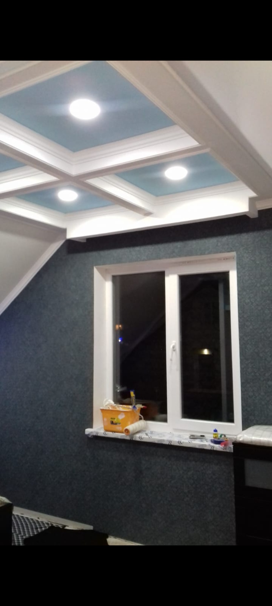 Wykończone wnętrze pokoju z dekoracyjnym podwieszanym sufitem z niebieskimi panelami i białymi listwami, ściany pokryte ciemną tapetą, na parapecie okiennym wiadro z farbą i wałek.