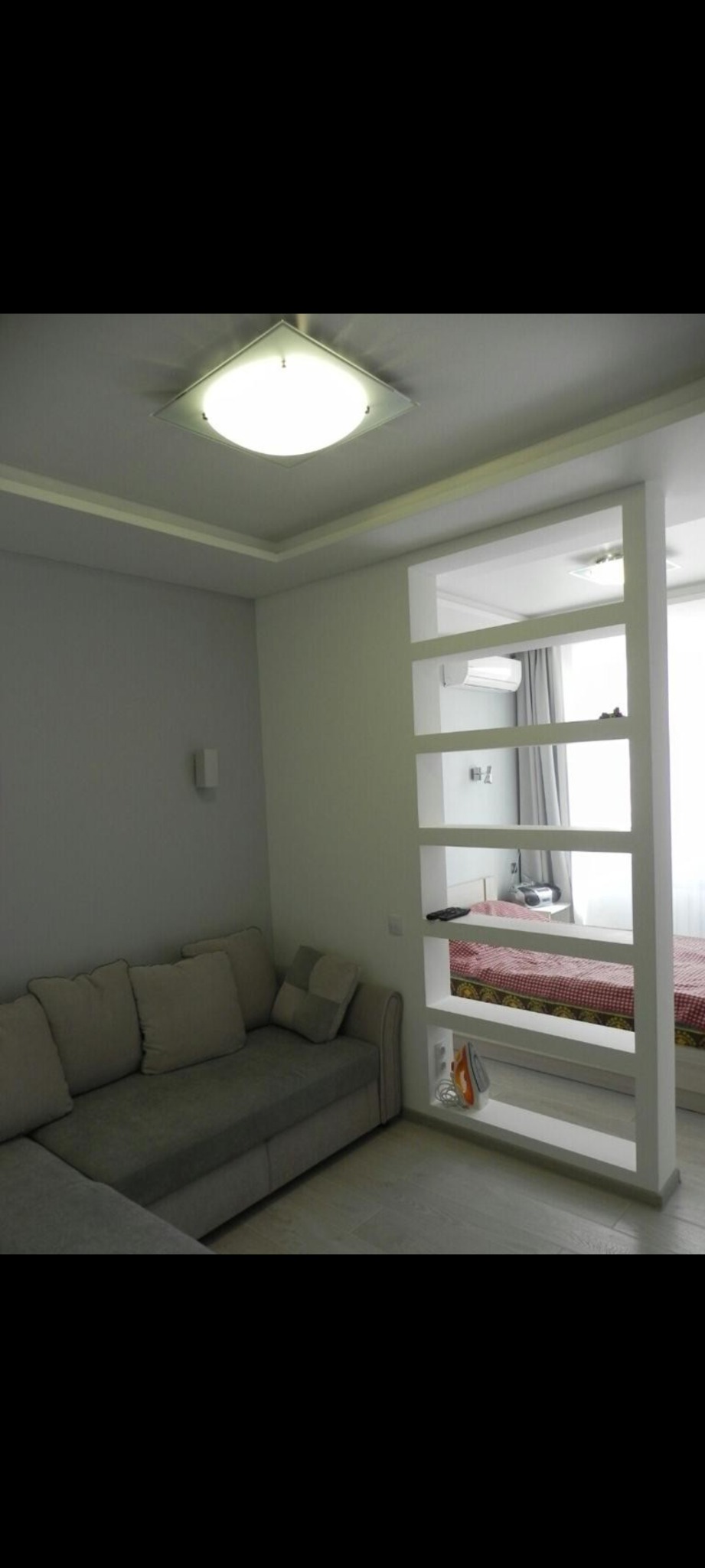 Nowoczesny salon z szarą sofą narożną i białą, ażurową ścianką działową z półkami, przez którą widać sypialnię z łóżkiem w kratę.