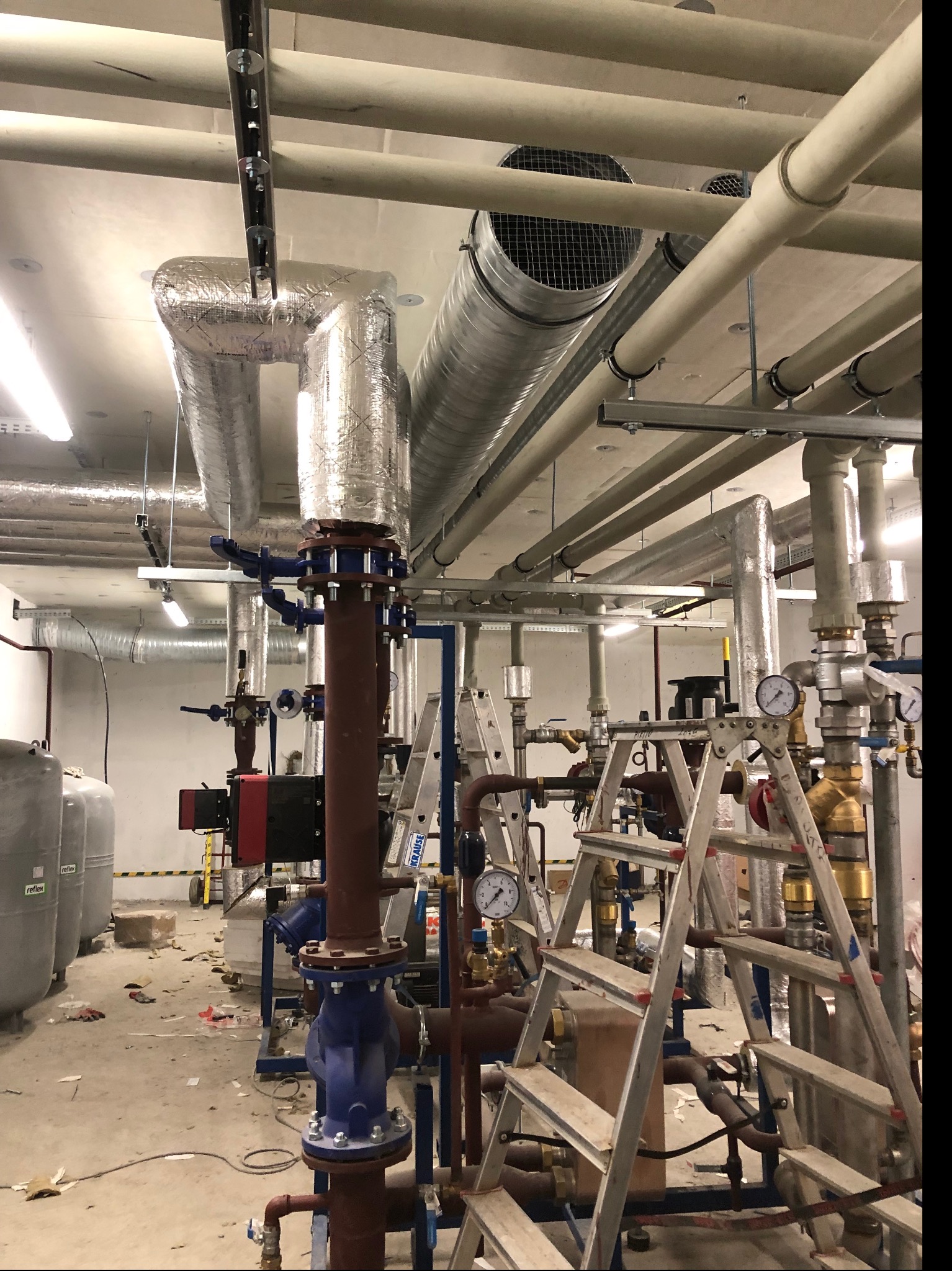Rozbudowana instalacja hydrauliczna w kotłowni, z widocznymi rurami, zaworami, manometrami, izolacją termiczną i drabinkami monterskimi, w trakcie montażu lub serwisu.
