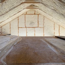 Thermo System - Poddasze z drewnianą konstrukcją pokryte grubą warstwą białej piany izolacyjnej, widoczne fragmenty podłogi zabezpieczone folią.