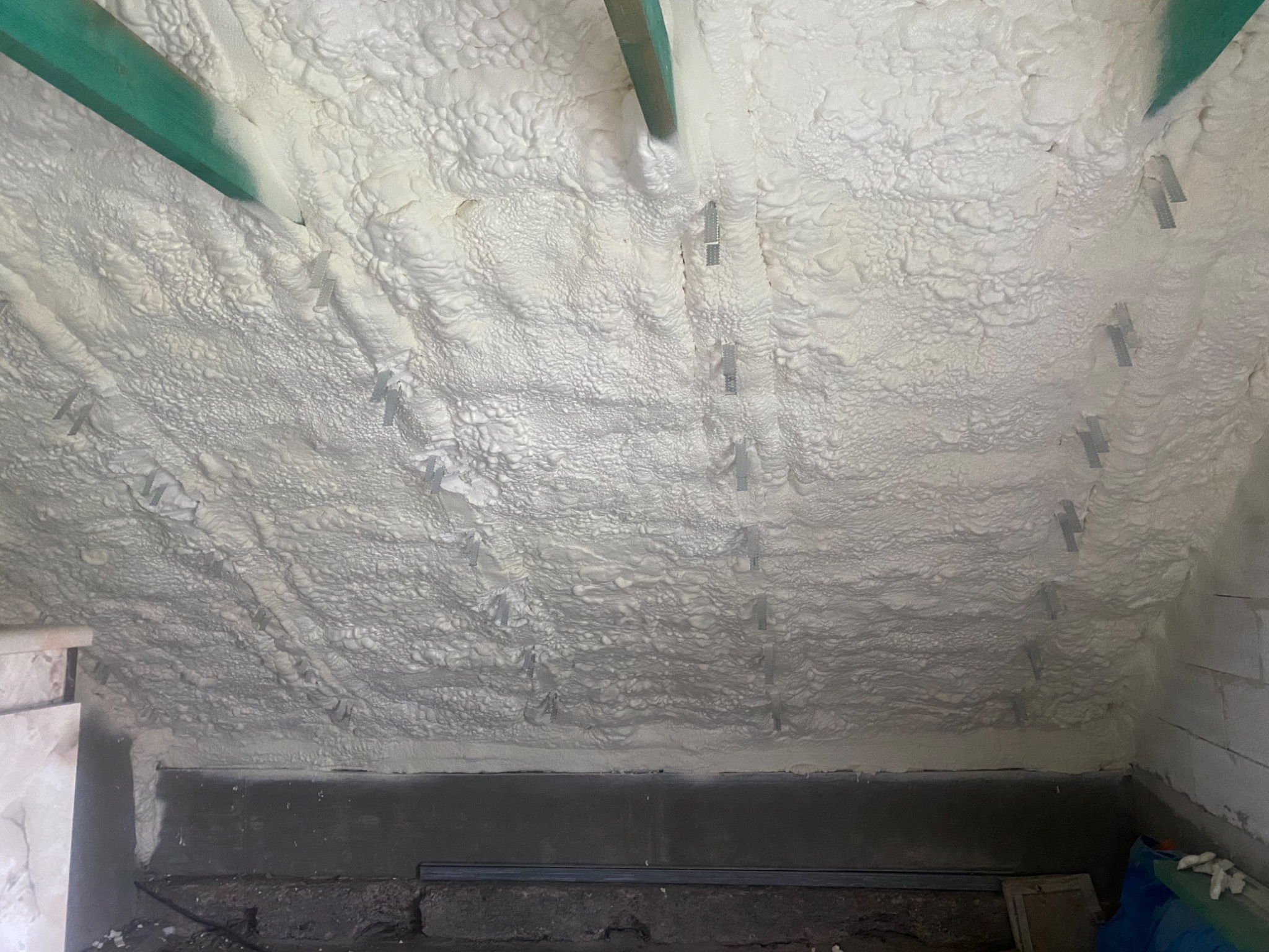 Poddasze ocieplone pianką PUR, widoczne metalowe elementy mocujące oraz drewniana belka stropowa w kolorze zielonym.