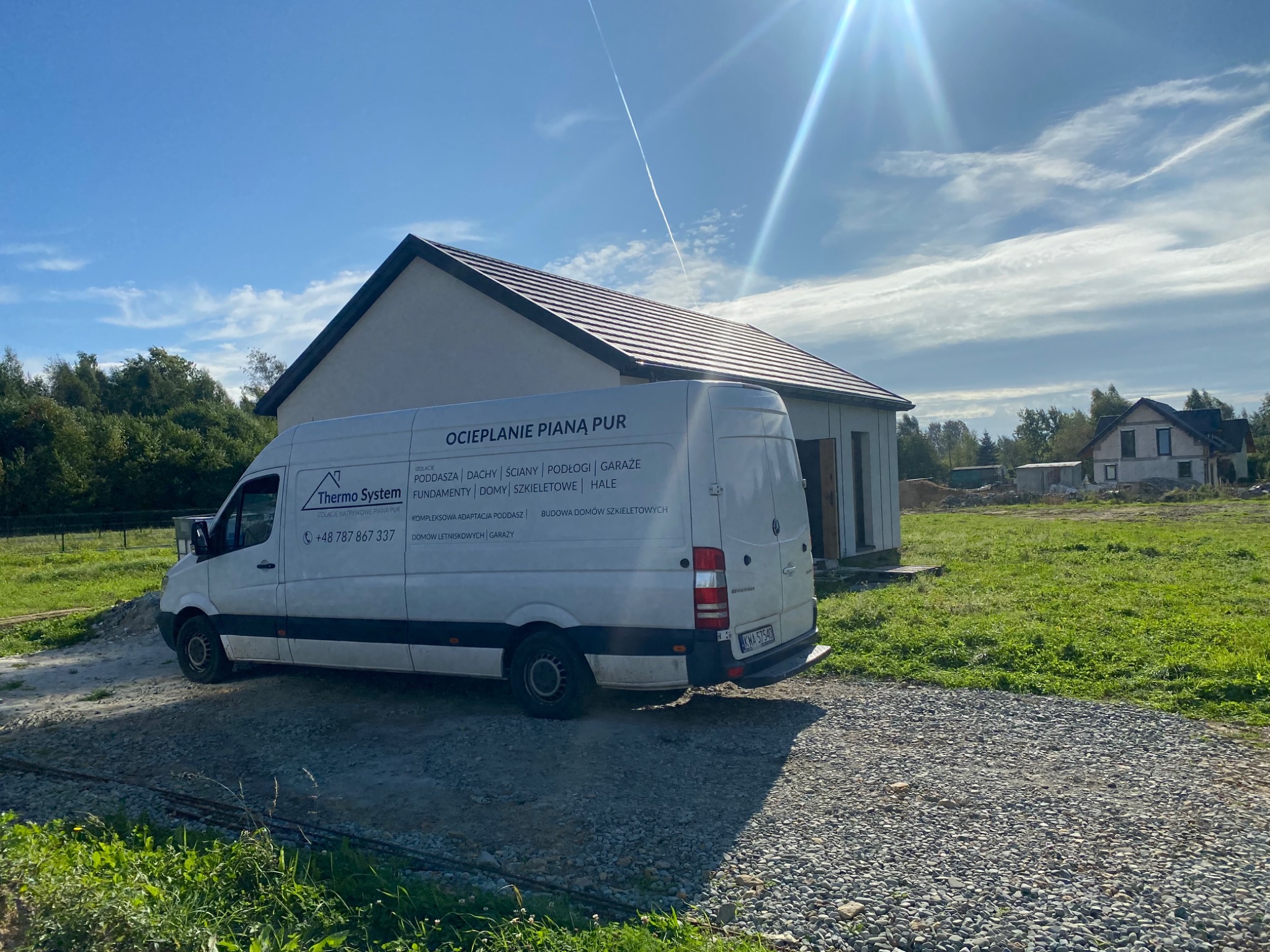 Biały samochód dostawczy z napisem 'Ocieplanie Pianą PUR' na tle budowanego domu jednorodzinnego w słoneczny dzień.